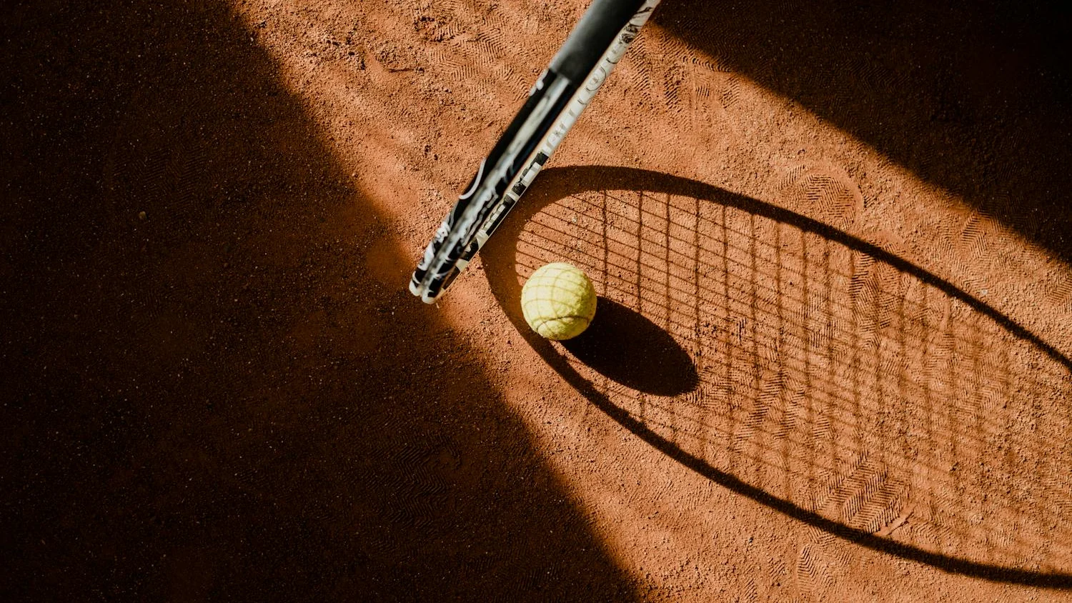 Tennisschläger und Ball auf einem Sandplatz im Abendlicht.